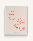 An art print titled "La Playa" featuring beach chairs, an umbrella, and the text "on a beach in Spain" in red, on a white background.