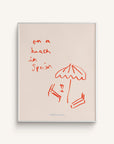 An art print titled "La Playa" featuring beach chairs, an umbrella, and the text "on a beach in Spain" in red, on a white background.